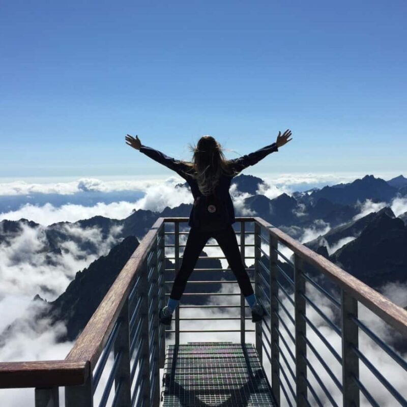 Woman looks out over a mountain range on a bridge with arms raised showing how integrated recovery has changed her life.